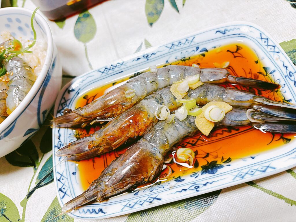 青い模様の角皿に並べられた、自家製の「天使の海老」のカンジャンセウ。醤油ベースのタレに、スライスしたにんにくや生姜、刻みネギが添えられ、一晩寝かせて味が染み込んだ艶やかな海老の様子。