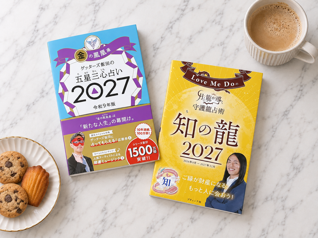 買ってきたゲッターズ飯田の五星三心占い2027 金の鳳凰座と、Love Me Doの守護龍占術 知の龍2027を、コーヒーとクッキーと一緒に並べた様子